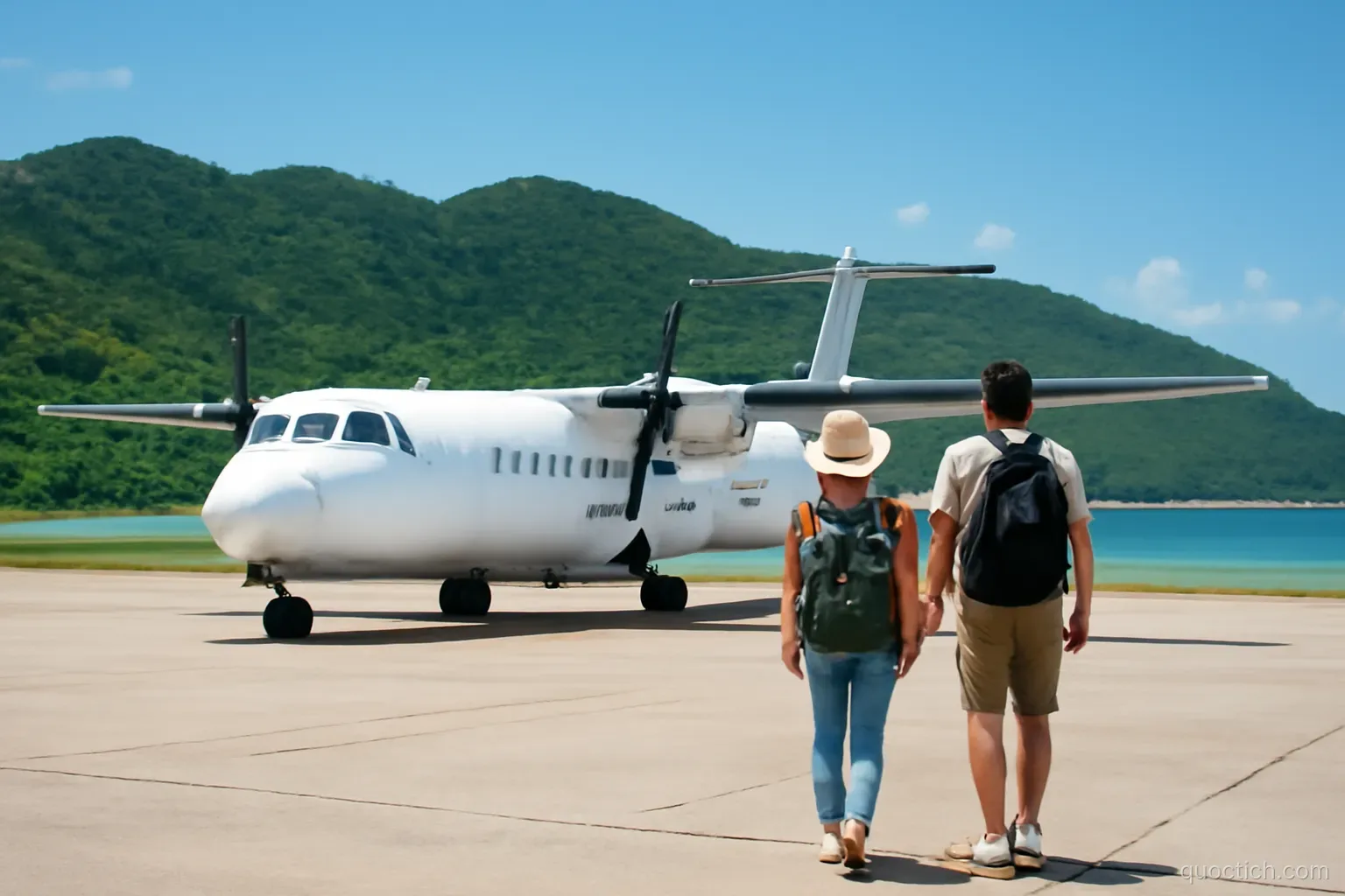 Small white turboprop plane parked on a sunny tropical airstrip with green hills and turquoise sea behind, a Vietnamese couple walking toward it with backpacks, wide shot